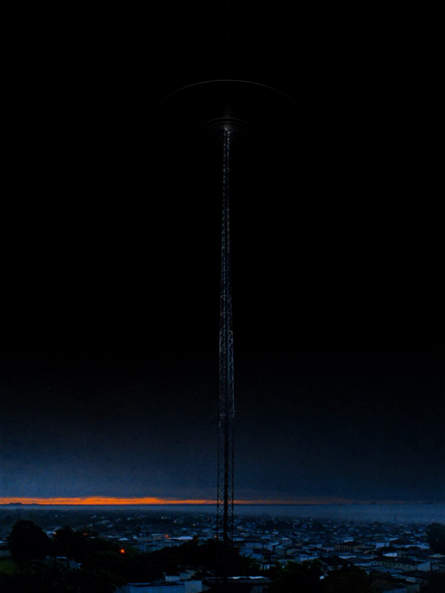 Torre de transmissão de rádio silhuetada contra o céu antes do amanhecer, com uma cidade pequena iluminada ao fundo